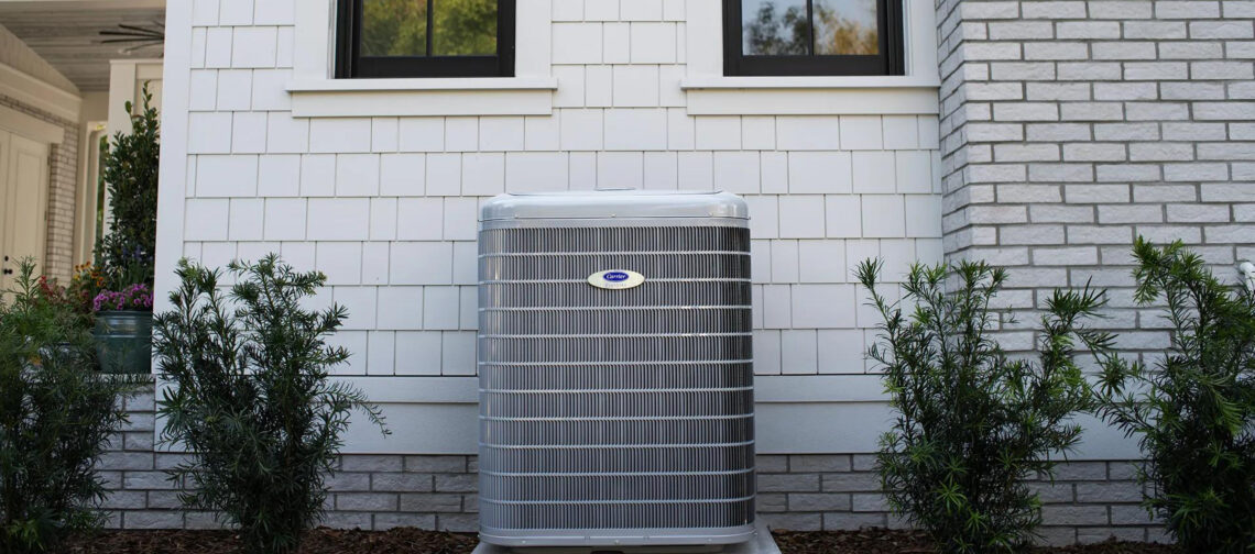 A carrier heat pump outside a white house with plants on both sides.